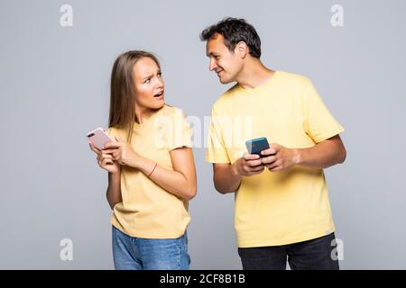 Portrait of couple with smart phones in hands, cheerful charming woman checking her email, man follows her secret correspondence with jealous expressi Stock Photo