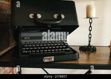 Vintage typewriter in black suitcase with metal lock put on wooden table near chain shaped candlestick and candle above in daylight Stock Photo