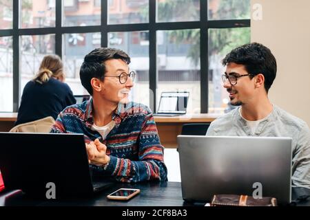 Positive coworkers in casual clothes and eyeglasses sitting at desk and working on project on laptops during workday in modern office Stock Photo