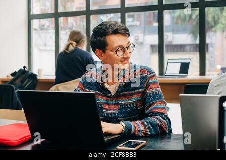 Young concentrated male in casual outfit and eyeglasses sitting at desk and typing on laptop while working on project in creative workspace Stock Photo