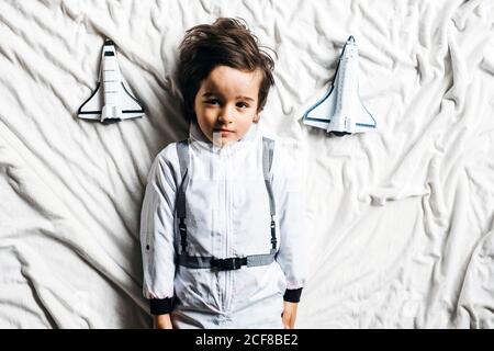 Top view of child wearing spacesuit lying on bed with toy spaceships and looking at camera Stock Photo