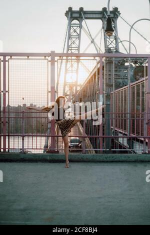 Side view of graceful female dancer in skirt balancing on tiptoe with outstretched arms while practicing choreographic elements on background of cityscape Stock Photo