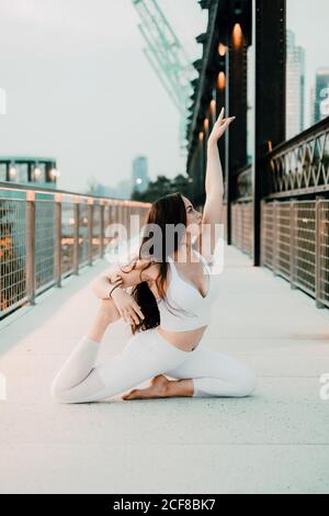 Side view of flexible female in sportswear exercising yoga in king pigeon pose while sitting on concrete bridge and practicing mindfulness Stock Photo