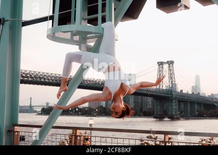 Low angle of tranquil female in sportswear hanging upside down with closed eyes while meditating on background of cityscape Stock Photo