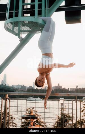 Low angle of tranquil female in sportswear hanging upside down with closed eyes while meditating on background of cityscape Stock Photo