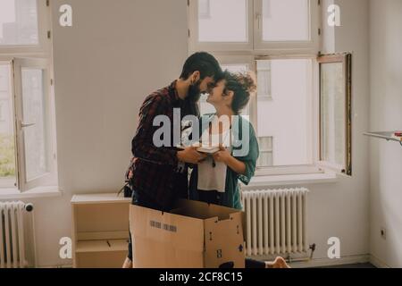 Hipster couple unpacking together boxes while standing barefoot in light room and cuddling Stock Photo