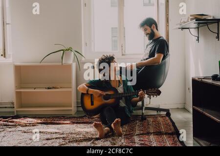 Happy Woman playing guitar and sitting on floor while man reading book and listening Stock Photo