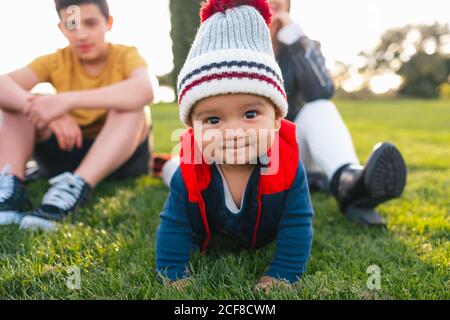 Cute ethnic toddler in warm clothes smiling and looking at camera while crawling on green meadow during family weekend in countryside Stock Photo