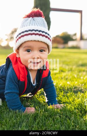 Cute ethnic toddler in warm clothes smiling and looking at camera while crawling on green meadow during family weekend in countryside Stock Photo
