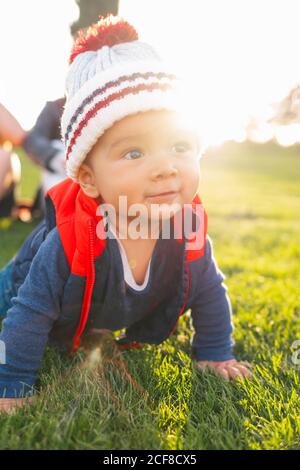 Cute ethnic toddler in warm clothes smiling while crawling on green meadow during family weekend in countryside Stock Photo