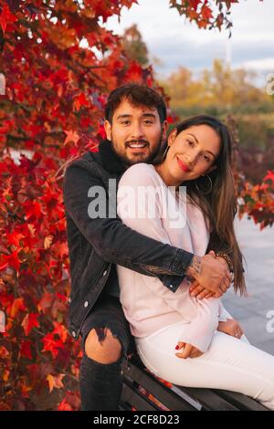 Happy young man and Woman embracing while sitting on bench near colorful autumn tree in park Stock Photo