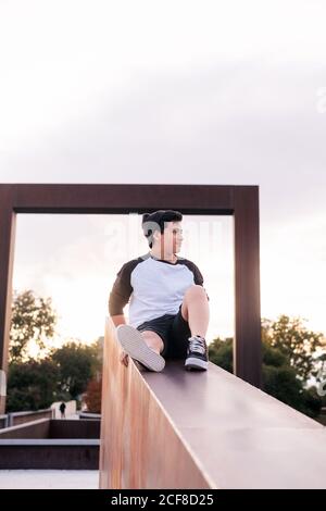Low angle of positive young man in casual wear sitting on stone fence and looking away while resting in city park in sunny evening Stock Photo