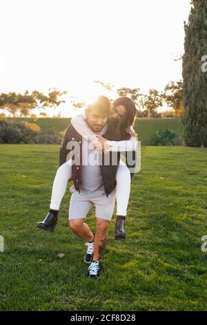 Cheerful young man giving piggyback ride to happy wife while enjoying sunny autumn day on green meadow in countryside Stock Photo