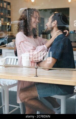 Side view of cheerful couple hugging in coffee shop drinking hot beverage and looking at each other Stock Photo