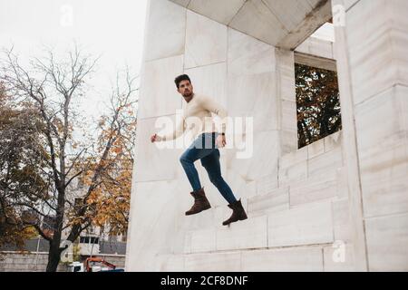 Side view of trendy young man in jeans and white sweater running in air by white building Stock Photo