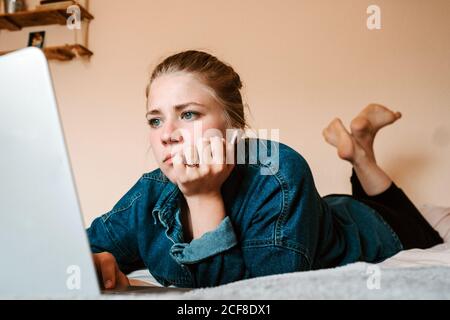 Thoughtful focused female in true wireless earbuds and casual clothes lying down on bed using laptop against beige wall in light apartment Stock Photo
