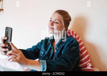 Joyful young female in wireless earphones and denim jacket smiling at screen and taking selfie with smartphone while relaxing on bed in modern apartment Stock Photo