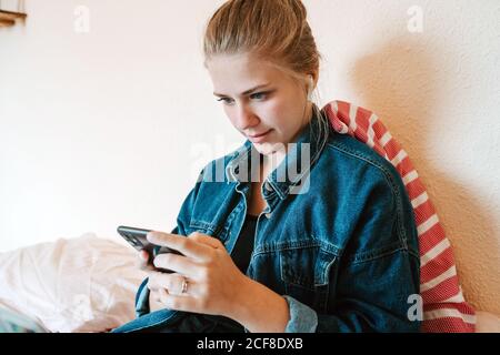 Young female in wireless earphones and denim jacket using smartphone while relaxing on bed in modern apartment Stock Photo