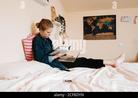 Serious pensive female student in casual clothes reading textbook with interest while sitting with legs crossed on bed and using laptop against modern interior of light cozy apartment Stock Photo