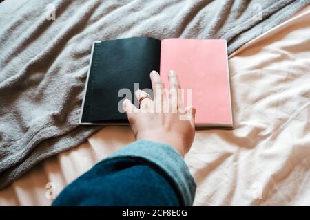 From above faceless female in casual clothes taking opened notebook with empty black and pink pages from soft bed in light modern bedroom Stock Photo