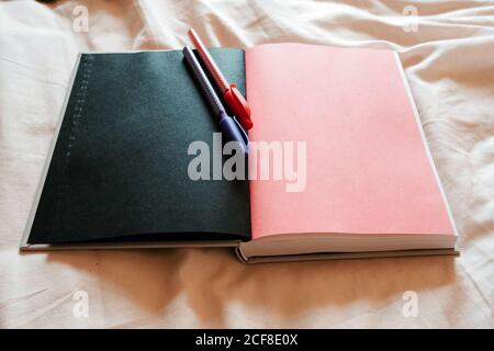 From above opened notebook with blank black and pink pages in composition with red and blue pens on white duvet on bed in light bedroom of modern apartment Stock Photo