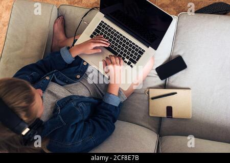 young female freelancer in casual wear with wireless headphones sitting on sofa and using laptop while working on remote project at home Stock Photo