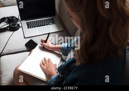 young female in casual outfit taking notes in diary while sitting on sofa with gadgets in living room Stock Photo