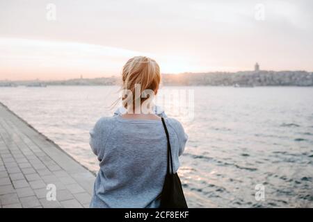 Back view of anonymous female traveler in casual outfit standing on embankment and admiring view of colorful amazing sunset Stock Photo