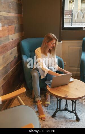 Young blonde happy female freelancer in casual clothes having tasty hot drink while sitting at round wooden table and using netbook in light modern cafe Stock Photo