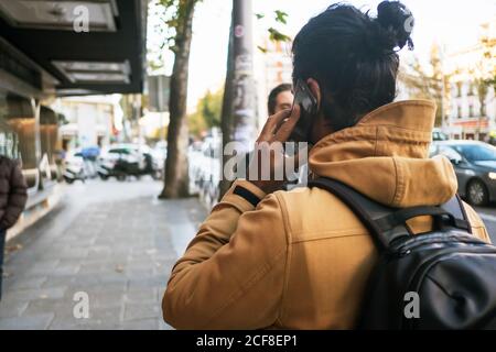 Back view of anonymous male in autumn jacket with long dark hair in bun talking on smartphone while walking down street in city with backpack Stock Photo