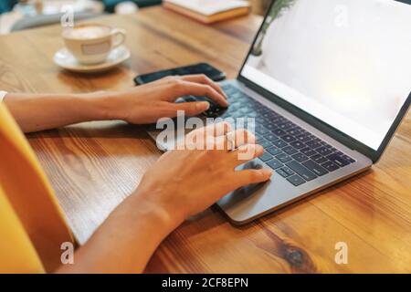 Cropped unrecognizable young female freelancer in casual clothes having tasty hot drink while sitting at wooden table and using laptop in light modern cafe Stock Photo