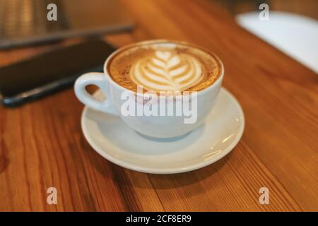High angle of white ceramic cup of yummy hot cappuccino covered with red lipstick marks on brown wooden table beside blurred smartphone in modern cafe Stock Photo