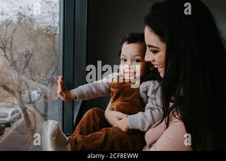 Side view of loving mother in casual clothes holding cute little son on hands while standing near window at home and pointing away Stock Photo