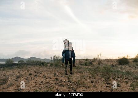 Full body man giving piggyback ride to happy girlfriend while standing in field against cloudy sundown sky in nature