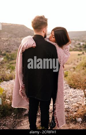 Back view of unrecognizable boyfriend hugging girlfriend covered with plaid while standing together on hill and admiring spectacular scenery on valley Stock Photo