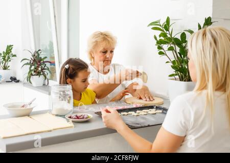 Little girl learning to roll dough and make homemade pastry or cookies with her mother and grandmother Stock Photo