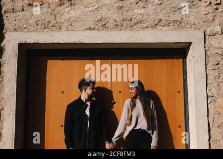 Smiling young ethnic couple in casual clothes holding hands and looking at each other with love while standing against entrance of aged stone building Stock Photo