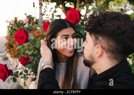 Smiling young ethnic couple in casual clothes looking at each other with love with roses in the background Stock Photo