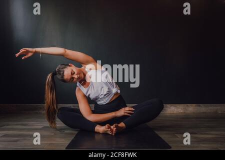 Young relaxed female with eyes closed and arm raised sitting in baddha konasana position on sports mat and practicing yoga in dark hall Stock Photo