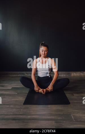 Diligent barefooted Woman in sportswear performing bound angle pose exercise on mat in light contemporary gym Stock Photo