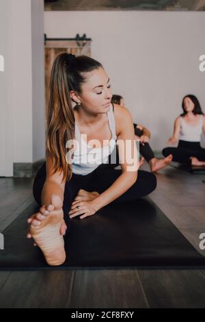 Diligent barefooted Woman in sportswear performing head to knee forward bend exercise on mat while other sporty people resting after training in light contemporary gym Stock Photo