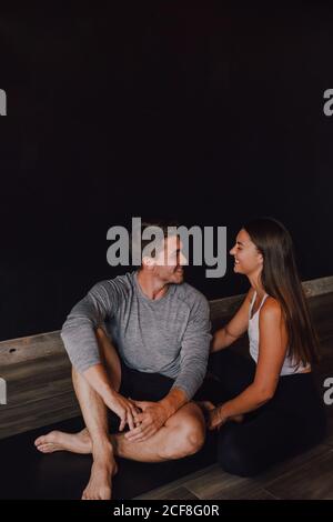 Magnificent slim Woman and happy adult man in sportswear embracing and smiling at each other while sitting on sports mat against black wall in contemporary yoga studio Stock Photo