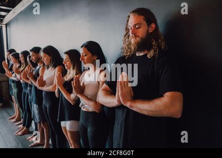 Group of happy young sporty people in activewear with eyes closed relaxing  and deep breathing while standing with namaste in tadasana and meditating  beside black wall in modern yoga studio stock photo