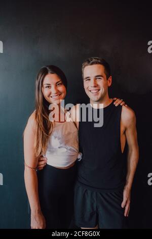 Attractive fit Woman and happy millennial man in sportswear embracing and smiling at camera while standing against black wall Stock Photo