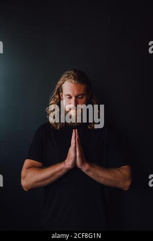 Handsome male athlete in sportswear with namaste praying hands performing yoga kakasana against black wall of contemporary workout room Stock Photo