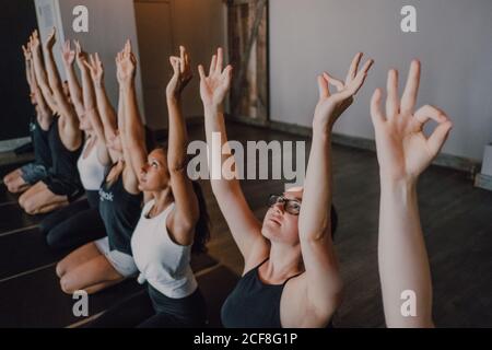 High angle of group of young sporty women and men in sportswear with arms raised and Gyan Mudra stretching body and meditating while sitting in virasana pose on sports mats and looking up training in modern yoga studio Stock Photo