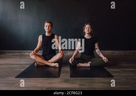 Calm young Woman and man in sportswear with eyes closed and legs crossed meditating together while sitting in padmasana position against back wall in contemporary yoga studio Stock Photo