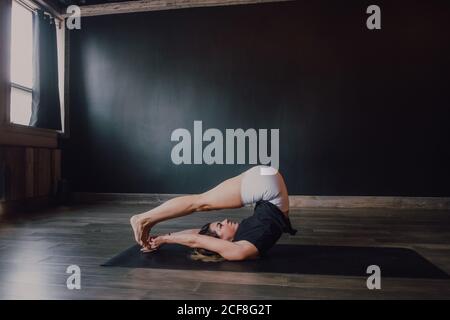 High angle side view of slender focused female athlete in sportswear stretching body in halasana position while training alone in spacious contemporary yoga studio Stock Photo