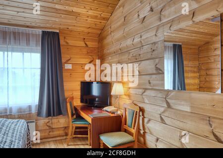 Cozy interior of country hotel room with mirror on wooden wall and TV on table with chairs in soft light of lamp Stock Photo