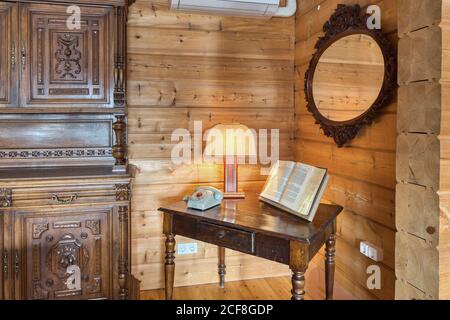 Table with open book arranged with lamp and telephone in corner of old fashioned interior with mirror on wooden wall and cupboard Stock Photo